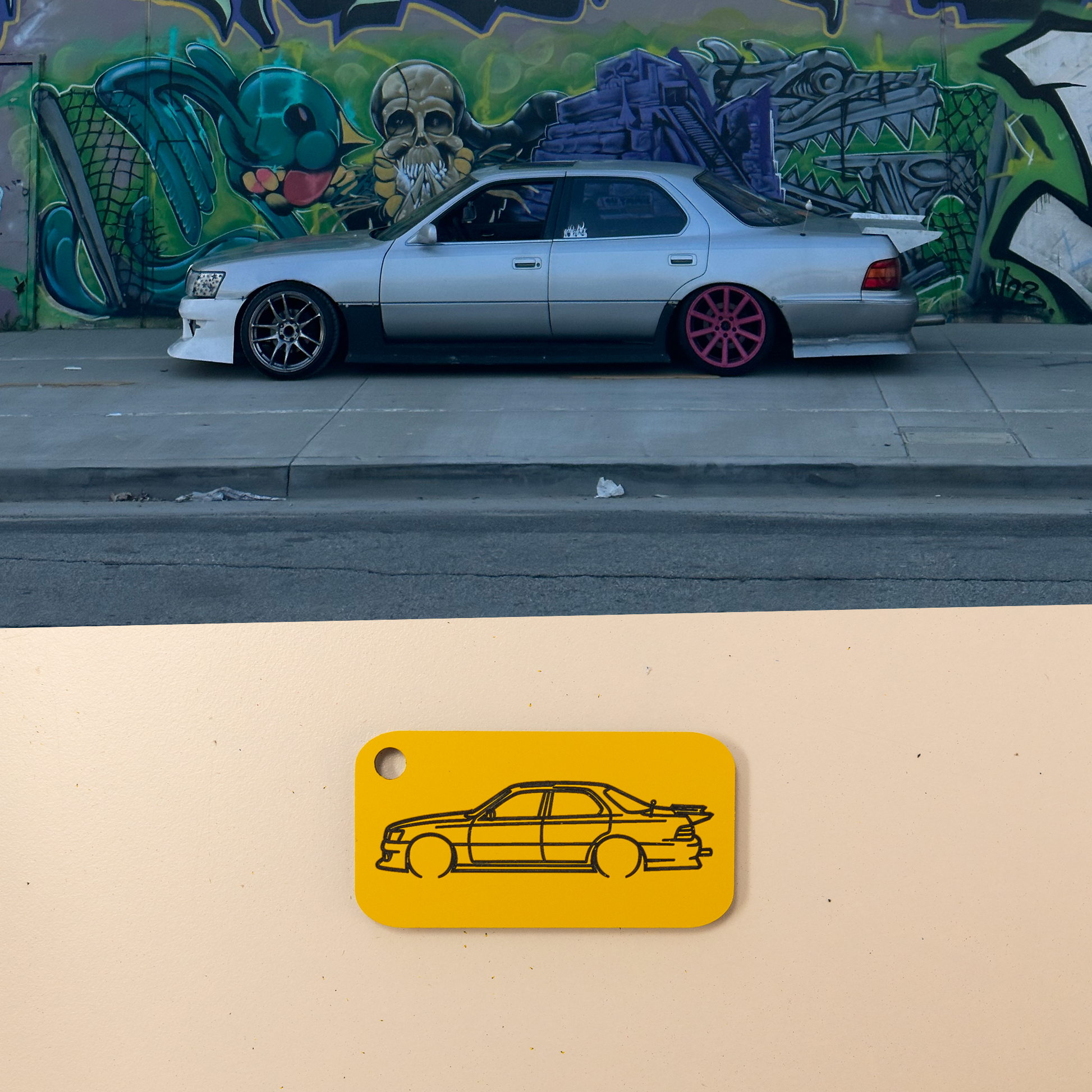 Silver Lexus LS400 with pink rims parked on a street next to a mural, with a yellow car keychain illustration below.